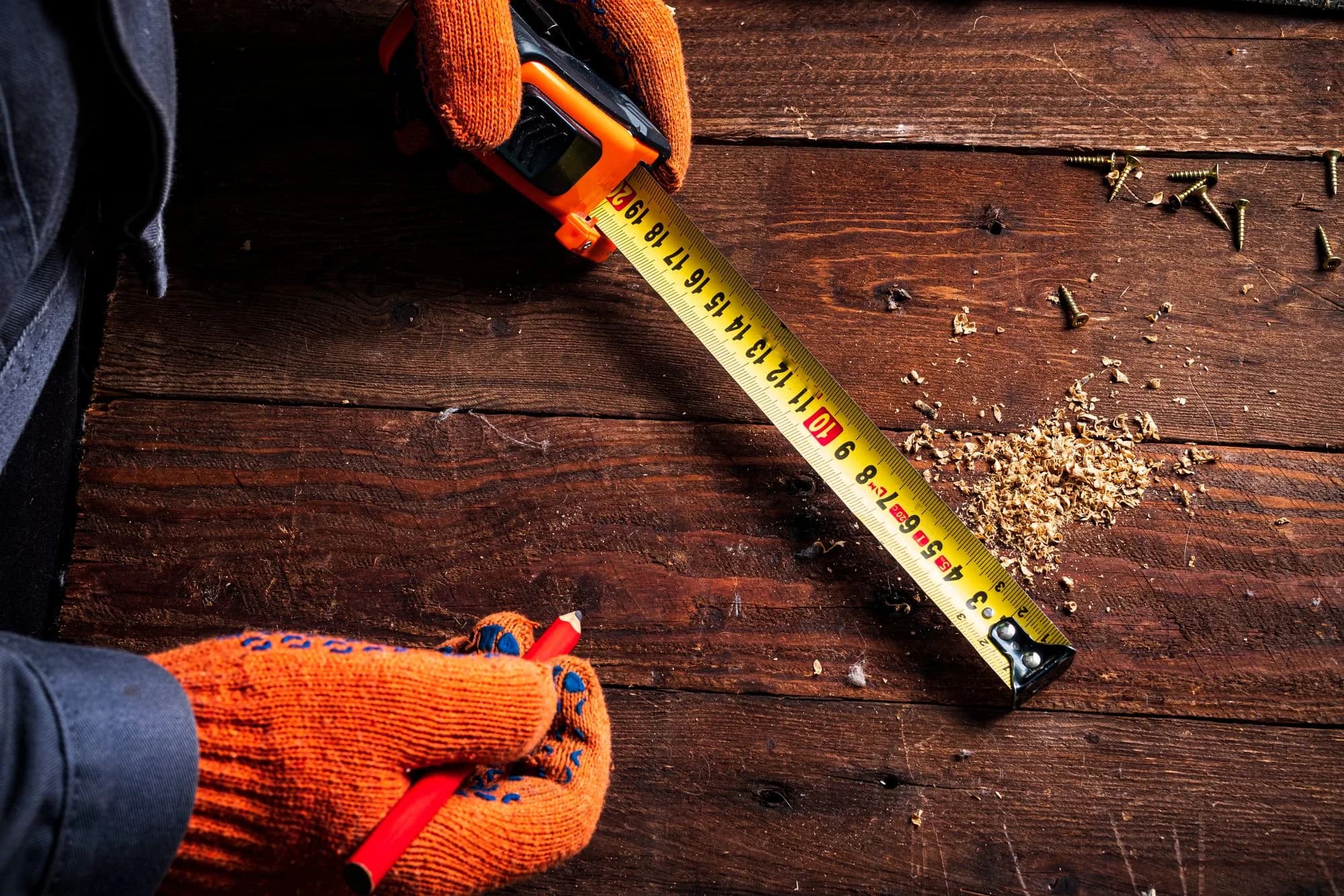 Gloved hands using a tape measure to check deck joist spacing on a wooden framing surface